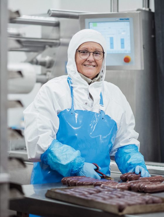 Collaboratrice de la branche Viandes d'Agrial. Elle porte une tenue de travail blanche avec un tablier bleu, des gants bleus et découpe les boudins noirs sur un plan de travail d'usine Collaboratrice de la branche Viandes d'Agrial. Elle porte une tenue de travail blanche avec un tablier bleu, des gants bleus et découpe les boudins noirs sur un plan de travail d'usine