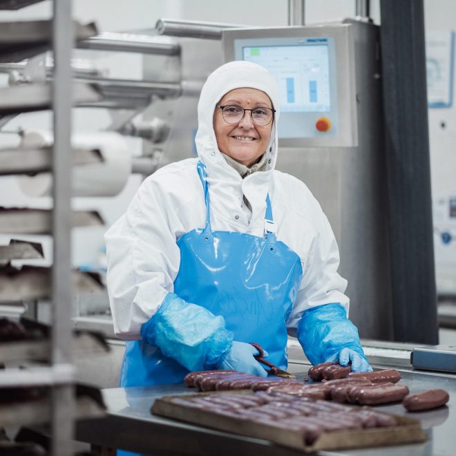Collaboratrice de la branche Viandes d'Agrial. Elle porte une tenue de travail blanche avec un tablier bleu, des gants bleus et découpe les boudins noirs sur un plan de travail d'usine Collaboratrice de la branche Viandes d'Agrial. Elle porte une tenue de travail blanche avec un tablier bleu, des gants bleus et découpe les boudins noirs sur un plan de travail d'usine